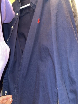 Mörkblå skjorta från Ralph Lauren - Klassisk mörkblå skjorta från Ralph Lauren med röd broderad logga på bröstet. Skjortan har långa ärmar, knappar framtill och är gjord i mjuk bomull. Perfekt till jeans eller chinos för en clean och tidlös look.