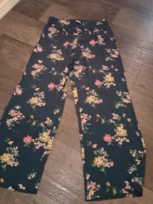 Blommiga vida byxor Jacqueline de Yong - Mörkgröna byxor från Jacqueline de Yong med blommigt mönster i rosa, gult och grönt. Vida ben och mjukt, lätt material som känns skönt mot huden. Perfekta för dig som gillar en avslappnad men ändå trendig look.