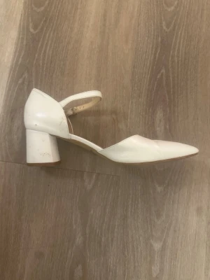 Vita pumps med blockklack från Bata - Säljer ett par stilrena vita pumps från Bata i storlek 38. Skorna har en spetsig tå, blockklack och justerbar rem runt ankeln. Tillverkade i slätt syntetmaterial med en låg klack som ger både höjd och komfort. Perfekta för dig som vill ha en clean och klassisk look. Världens bästa bröllop sko. Bar dessa utan problem under dag och natt 