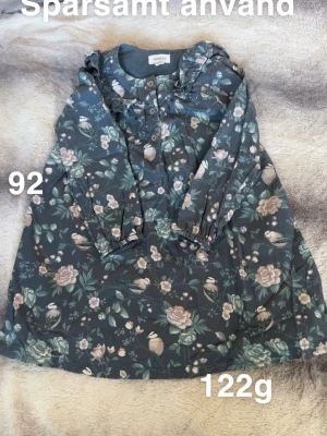 Blommig långärmad klänning Newbie - Supersöt klänning från Newbie i mörk botten med blommigt mönster och fåglar. Långärmad modell med volangdetaljer vid axlarna och knappar framtill. Perfekt för dig som gillar romantisk stil.🌸Sparsamt använd, storlek 92 