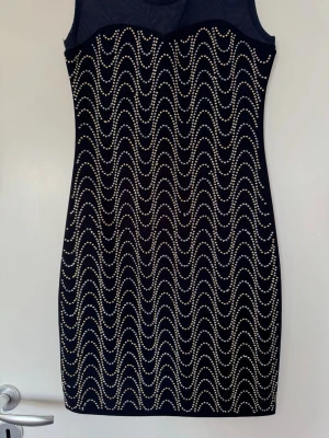 Svart kort klänning med pärldetaljer - Snygg svart kort klänning med transparent mesh upptill och vågformade pärldetaljer framtill. Ärmlös modell med figurnära passform, perfekt för dig som vill sticka ut. Materialet känns stretchigt och bekvämt.