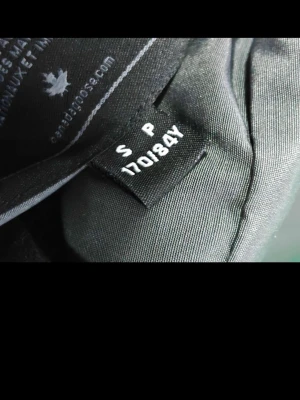 Grå dunjacka från Canada Goose S - Säljer en grå dunjacka från Canada Goose i storlek S. Jackan har huva och medföljande avtagbar pälsdetalj, samt ribbade muddar vid ärmsluten. Klassisk patch på ärmen och stängs med både knappar och dragkedja. Perfekt för kalla dagar. Jackan Har NFC 