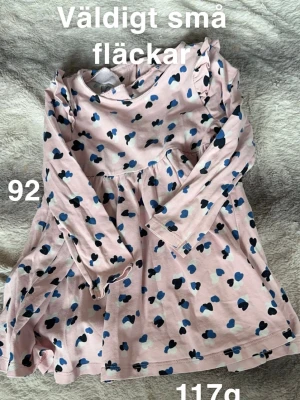 Rosa klänning med hjärtan, stl 92 - Söt rosa klänning med lång ärm och hjärtmönster i blått, svart och vitt. Klänningen har en rund hals och lätt utställd kjol, perfekt för dig som gillar gulliga detaljer. 🌸 Små fläckar. Storlek 92