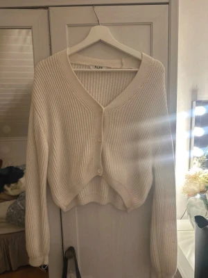 Beige ribbad kofta från NA-KD - Mysig beige kofta från NA-KD med ribbad struktur och v-ringning. Koftan har knappar framtill, lång ärm och en något croppad passform som ger en chill vibe. Perfekt att slänga över ett linne eller t-shirt under kyliga dagar.