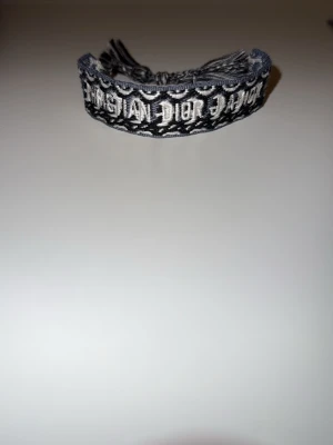 Christian Dior armband - Exklusivt Christian Dior J’Adior-armband i elegant svart och grå färgskala. Armbandet är tillverkat i vävd textil med den välkända “CHRISTIAN DIOR J’ADIOR” logotypen framtill, vilket ger en sofistikerad men samtidigt modern look. Justerbar knytning baktill med dekorativa tofsar för optimal passform och komfort.