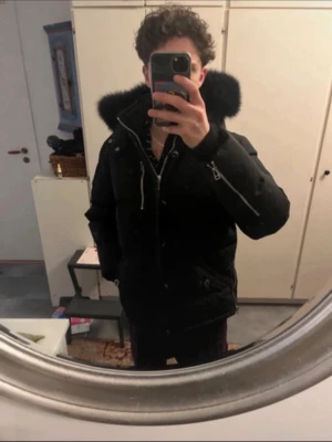 Svart Moose Knuckles dunjacka - Moose Knuckles svart dunjacka med stor pälskantad huva och silverfärgade detaljer. Jackan har flera dragkedjor, tryckknappar och logga på ärmen. Insidan är fodrad och jackan är perfekt för kalla dagar. Snygg, stilren och riktigt varm vinterjacka. Pris kan diskuteras.