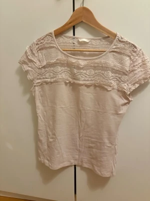 Ljusrosa blus med spetsdetaljer - Supersöt ljusrosa blus med romantiska spetsdetaljer över axlar och bröst. Perfekt för dig som gillar en feminin och luftig stil. Materialet känns mjukt och lätt. I väldigt bra skick! Skriv gärna vid funderingar!