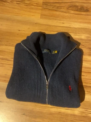 Marinblå stickad kofta Polo Ralph Lauren - Säljer en marinblå stickad kofta från Polo Ralph Lauren med ribbad struktur och hel dragkedja framtill. Klassisk röd logga broderad på bröstet och hög krage. Perfekt för dig som vill ha en stilren och tidlös look.