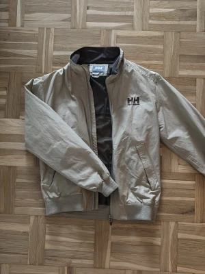 Beige vindjacka från Helly Hansen - Säljer en beige vindjacka från Helly Hansen med svart meshfoder och HH-logga på bröstet. Jackan har dragkedja, ribbade muddar och praktiska fickor. Perfekt för dig som vill ha en stilren och funktionell jacka till vardags.