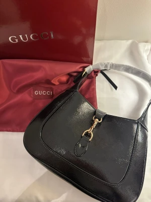 Svart axelväska. - Säljer en snygg svart axelväska i glansigt skinn med guldfärgade metalldetaljer och klassisk form. Väskan har en stilren design med dekorativ spänne framtill och kommer med original dustbag och box. Obs detta är inte orginel men är väldigt bra kvalite