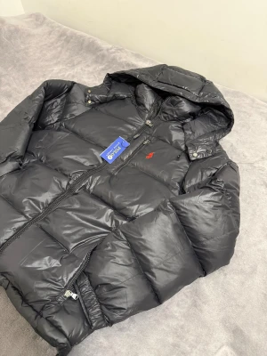 Ralph Lauren jacka  - Svart pufferjacka från Polo Ralph Lauren med huva, dragkedja och två sidofickor. Jackan har en röd broderad logga på bröstet och quiltad design. Perfekt för kalla dagar och har en snygg, modern look.