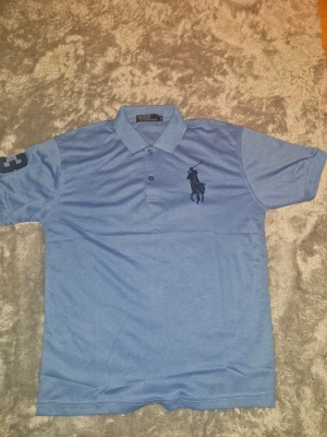 Sportpiké tröja från Polo Ralph Lauren - Säljer en klassisk blå pikétröja från Polo Ralph Lauren med stor mörkblå logga broderad på bröstet och siffran 3 på ärmen. Tröjan har krage, knappar framtill och är kortärmad. Perfekt till en avslappnad men ändå stilren look. Material: Polyester