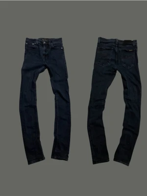 Nudie Jeans - Säljer ett par svarta jeans från Nudie med klassisk femficksdesign och snygga sömdetaljer på bakfickorna. Jeansen har normal midja och är tillverkade i stretchig denim för en bekväm passform.