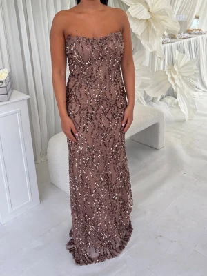 Brun glittrig långklänning - Säljer en brun axelbandslös långklänning med glittriga paljettdetaljer. Klänningen har en elegant och figurnära passform, perfekt för speciella tillfällen. Materialet känns lyxigt och skimrar snyggt i ljuset.