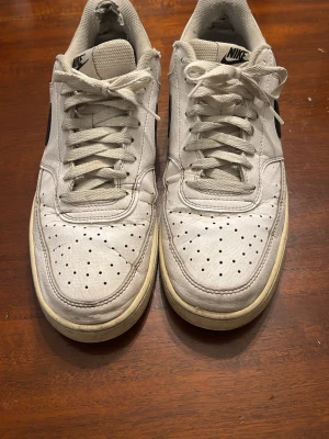 Nike Air Force 1 vita sneakers - Klassiska Nike Air Force 1 sneakers i vitt skinn med svart swoosh på sidan. Lågt skaft, perforerad tå och snörning framtill. Tjock, platt sula med cirkulärt mönster undertill. Perfekta för dig som gillar retrostil och streetwear.