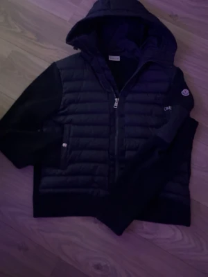 Svart pufferjacka från Moncler - Säljer en svart pufferjacka från Moncler med huva och ribbstickade ärmar. Jackan har quiltad framsida, dragkedja och Moncler-logga på ärmen. Perfekt för dig som vill ha en stilren och varm jacka med snygga detaljer.