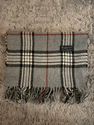 Grå rutig halsduk från Burberrys - Klassisk grå halsduk från Burberrys i mjuk lammull. Halsduken är i väldigt bra skick och endast använd ett par gånger! Hör av er vid frågor/eller för fler bilder! Pris går alltid att diskutera!