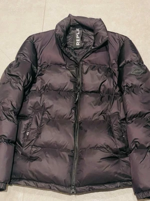 Replay jacka - Säljer en svart pufferjacka från Replay med hög krage och dragkedja framtill. Jackan har två stora fickor på sidorna och Replay-logga på ärmen. Perfekt för kalla dagar och riktigt skön att bära. Klassisk och stilren design som funkar till det mesta. Har kvitto+lapparna kvar