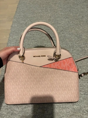 Michael Kors rosa handväska med logga - Säljer en snygg stor handväska från Michael Kors i ljusrosa och korall med MK-logga över hela väskan. Väskan har både handtag och justerbar axelrem med guldkedja, samt dragkedja upptill. Insidan har flera fack och dragkedja för smart förvaring. Väldigt fint skick. 