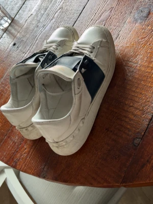 Valentino Garavani vita sneakers - Säljer ett par snygga vita Valentino Garavani sneakers med bred svart rem över mitten. Skorna har rund tå, platt sula och snörning. Tillverkade i skinn med klassisk design och diskreta nitar på sidan av sulan. Perfekta för dig som gillar stilrena och lyxiga sneakers.