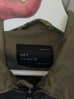 Olivgrön overshirt från Zara Man - Säljer en olivgrön overshirt från Zara Man i storlek S. Jackan har två bröstfickor med knappar, dragkedja framtill och coola broderade patches på ärmen. Tillverkad i bomull och perfekt för lager på lager. Enkel, stilren och med en avslappnad vibe.