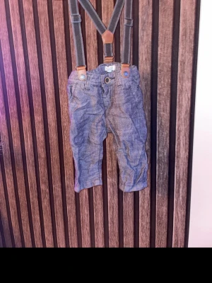 Newbie byxor med hängslen storlek 62 - Säljer ett par blå newbie byxor med snygga hängslen och knappdetalj i midjan. Byxorna har ett diskret rutigt mönster och är tillverkade i ett mjukt bomullsmaterial. Perfekt för dig som vill ha en klassisk men cool look. Storlek 62