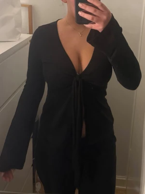 Svart öppen och v ringad jätte snygg topp  - Säljer en svart långärmad omlottopp med djup v-ringning och knyt framtill. Toppen har en croppad känsla och vida ärmar som ger en avslappnad men ändå edgy look. Perfekt att styla med höga jeans eller kjol för en trendig vibe.