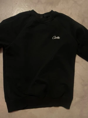 Svart sweatshirt från Crofts - En svart sweatshirt från Crofts med enkel och clean design. Tröjan har rund halsringning, långa ärmar och en liten vit logga broderad på bröstet. Perfekt för dig som gillar en stilren look och vill ha något bekvämt till vardags.