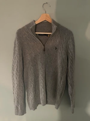 Grå kabelstickad tröja Polo Ralph Lauren - Kabelstickad grå tröja från Polo Ralph Lauren med halv dragkedja och klassisk broderad logga på bröstet. Tröjan har ribbade muddar, hög krage och är långärmad. Perfekt för kyliga dagar och riktigt snygg till jeans. Har en liten sydd del som knappt syns.