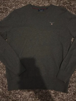 Mörkbrun stickad tröja från Gant - Säljer en mörkbrun stickad tröja från Gant med rund halsringning och diskret logga broderad på bröstet. Tröjan har långa ärmar och ribbade muddar vid ärmslut och nederkant. Perfekt för dig som gillar klassisk och enkel stil.