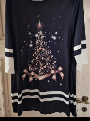 Svart julklänning med julgranstryck - Svart långärmad klänning med vitt och champagnefärgat julgranstryck framtill, dekorerad med stjärnor och rosetter. Klänningen har vita detaljer på ärmar och nederkant samt rund halsringning. Perfekt för vinterns festligheter.