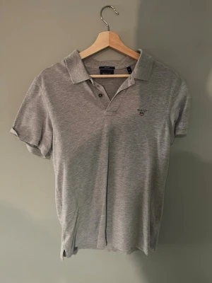 Grå pikétröja från Gant - Säljer en klassisk grå pikétröja från Gant med korta ärmar och liten logga broderad på bröstet. Tröjan har krage och två knappar framtill. Perfekt till jeans eller shorts för en clean och enkel look.