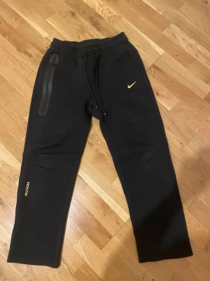 Svarta NOCTA x Nike mjukisbyxor - Svarta mjukisbyxor från Nike och NOCTA med gul logga på benet och klassisk Nike Swoosh. Byxorna har dragsko i midjan och en snygg dragkedjeförsedd ficka på sidan. Perfekta för chill eller streetwear-stil. Storlek M det kan även passa storelk S.