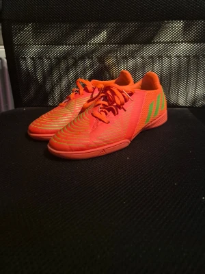 Orangea adidas fotbollsskor med snörning - Säljer ett par riktigt snygga orangea adidas  innefotbollsskor med gröna detaljer och klassiska ränder på sidan. Skorna har snörning och är tillverkade i syntetmaterial med meshpartier för bra ventilation. Perfekt när man börjar bli varm! Kan tvättas innan de skickas!