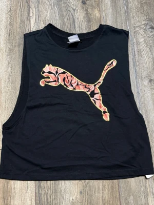 Svart croppad linnetopp Puma Kenza - Säljer en svart croppad linnetopp från Puma x Kenza-kollektionen. Toppen har ett stort Puma-tryck framtill med rosa och svart mönster. Ärmlös modell med breda ärmhål och rund halsringning. Perfekt till jeans eller shorts!