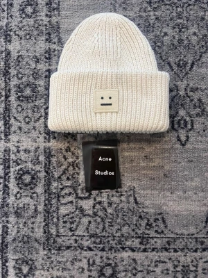 Vit stickad mössa från Acne Studios - Säljer en vit stickad mössa från Acne Studios med det klassiska ansiktsloggan framtill. Mössan är ribbstickad och har en uppvikt kant för extra stilpoäng. Perfekt accessoar för att hålla värmen och se cool ut under kyliga dagar.