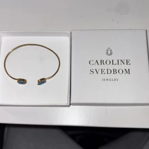 Guldarmband med blå stenar Caroline Svedbom - Snyggt och stilrent guldarmband från Caroline Svedbom med två gnistrande blå stenar i oval form på öppna ändarna. Perfekt accessoar för att lyfta din outfit med lite färg och elegans. Kommer i original ask. Nypris 799