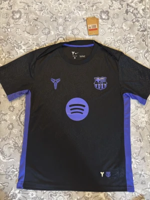 Barcelona X Kobe  - Säljer en svart FC Barcelona x Nike tröja med lila detaljer och loggor. Tröjan har ett lila Spotify-tryck på bröstet, Kobe Bryant-symbolen och FCB-emblemet i lila. Snygga lila paneler längs sidorna och ett subtilt mönster över hela tyget. Perfekt för dig som älskar fotboll och streetwear.