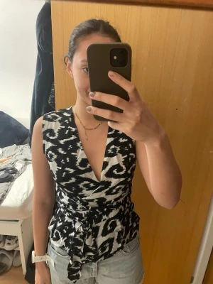 Svartvit omlottopp från DAY - Säljer en svartvit omlottopp från DAY med coolt mönster och långt knyte i midjan som man kan knyta hur man vill. Så fin till fest eller middag, storlek 38= S/M