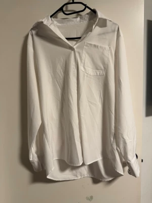 Vit oversized skjorta med ficka - Klassisk vit skjorta i oversized modell med lång ärm och bröstficka. Skjortan har knappar framtill och en avslappnad passform som ger en cool vibe. Perfekt att styla med jeans eller kjol för en clean look.