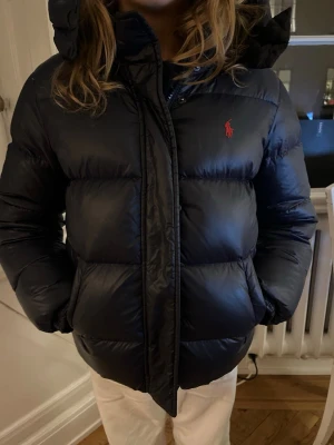 Svart pufferjacka från Ralph Lauren - Säljer en svart pufferjacka från Ralph Lauren med huva och dragkedja. Jackan har en klassisk röd logga på bröstet och två fickor med dragkedja. Perfekt för kalla dagar och riktigt skön att bära. Passar dig som vill ha en stilren och varm jacka.