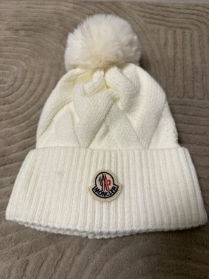 Vit stickad Moncler mössa med tofs - Säljer en vit stickad mössa från Moncler med stor fluffig tofs upptill och klassisk Moncler-logga framtill. Mössan har bred ribbad kant och ett snyggt flätmönster. Perfekt accessoar för kalla dagar.