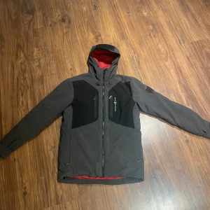 Svart och grå vindjacka från sail racing - Säljer en svart och grå vindjacka från sail racing med huva och rödfärgat innerfoder. Jackan har dragkedja framtill, två bröstfickor med dragkedja och svarta paneler på bröstet och ryggen. Perfekt för dig som gillar stilren och funktionell design.
