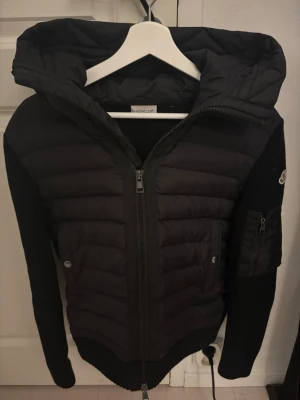 Svart Moncler pufferjacka med huva - Svart pufferjacka från Moncler med stickade ärmar och huva. Jackan har dragkedja framtill, ribbade muddar och en ficka med dragkedja på ärmen. Ikonisk Moncler-logga på vänster ärm och snygga detaljer som metallknappar. Perfekt för kyliga dagar.