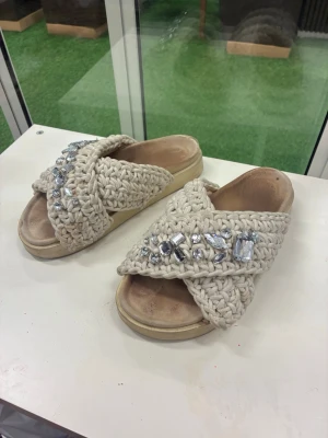 Beige sandaler med stenar från Inuikii - Säljers ett par chunky sandaler från Inuikii i beige stickad design med stora glittriga stenar på ovandelen. Skorna har korslagda band, öppen tå och platt sula. Perfekta för sommaren och ger en cool boho-vibe till din outfit.