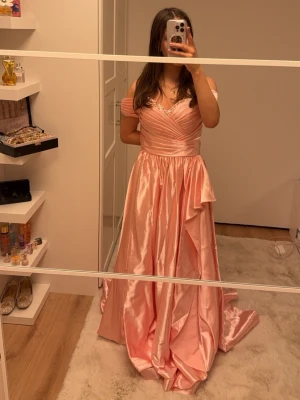 Ljusrosa långklänning i satin - Säljer en drömmig ljusrosa långklänning i glansigt satinmaterial. Klänningen har offshoulder-ärmar, v-ringad omlottdesign upptill med pärldetaljer vid urringningen och en snygg slits framtill. Kjolen är vid och faller elegant mot golvet. Jag är 164cm och den är lite för lång för mig så är man längre skulle den passa jättefint annars går det bra att sy upp. Klänningen är oanvänd och har inga defekter. Ett tips är även att lämna in klänningen till en kemtvätt så man får bort det skrynkliga. 