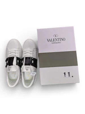 Valentino Garavani Rockstud sneakers - Säljer ett par vita Valentino Garavani Rockstud sneakers i skinn med svart band och nitar över vristen. Klassisk låg modell med rund tå, snörning och coola pyramidformade detaljer runt sulan. Perfekta för dig som vill ha något extra till din outfit.
