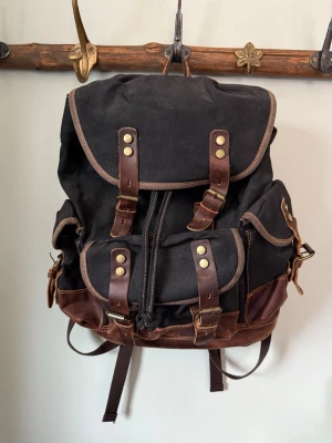 Svart och brun ryggsäck med läderdetaljer - Retro ryggsäck för herr, uppskattad av damer, perfekt som unisex. Tillverkad i äkta koskinn och oljevaxad canvas, otroligt slitstark och blir snyggare ju mer den används. Stilren vintagekänsla, perfekt för vardag, resor, arbete och outdoor, med plats för laptop/notebook. Helt ny i oöppnad förpackning.  -Den på bild som inte är i förpackning är ett visnings exemplar från vår förra butik. Har två st kvar en i brunt också. Ny pris på Amazon 3030kr    Ryggsäcksstorlek är L32 x B15 x H45 cm