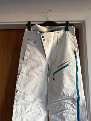  Peak Performance Gore-Tex skalbyxor  - Säljer ett par vita täckbyxor från Peak Performance med Gore-Tex-material. Byxorna har blå dragkedjor, flera fickor och en clean look. Perfekta för vintern och skidåkning, med slitstarka detaljer och snygg design.