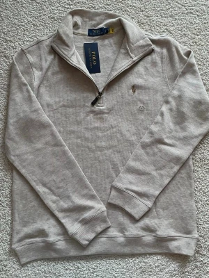 Beige half zip tröja Polo Ralph Lauren - Säljer en stilren beige tröja från Polo Ralph Lauren med klassisk broderad logga på bröstet. Tröjan har half zip-dragkedja, ribbade muddar och är tillverkad i mjuk bomull. Perfekt för dig som gillar en clean och avslappnad look. Tveka inte att höra av er vid frågor eller funderingar🤗